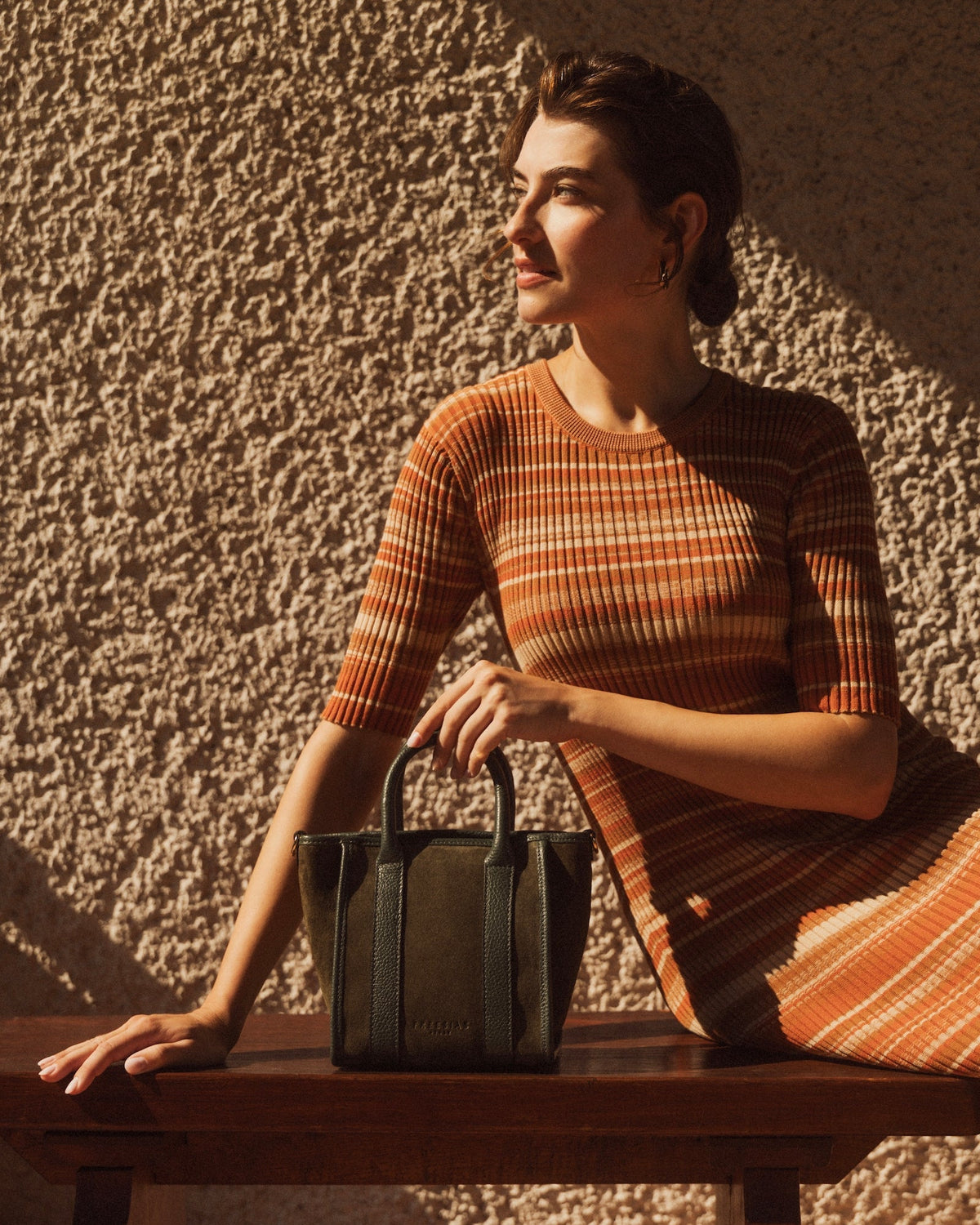 A woman in a rust-orange and brown striped knit dress sits on a wooden bench against a textured stucco wall, holding a structured dark green leather handbag.