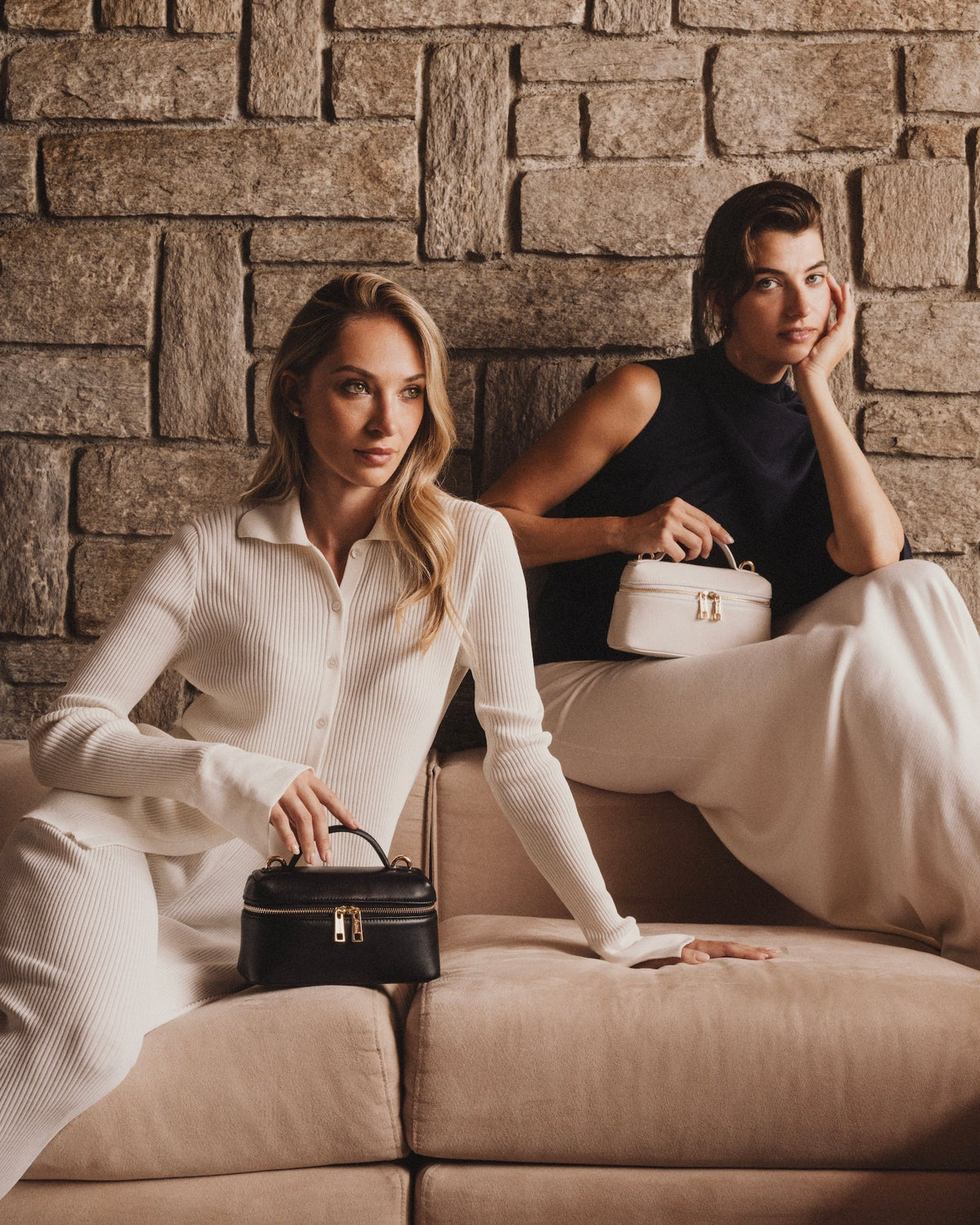 Two women posing on a beige sofa against a rustic stone wall, each holding a small structured handbag. The woman in the foreground wears a white ribbed long-sleeve top and pants, holding a black handbag.