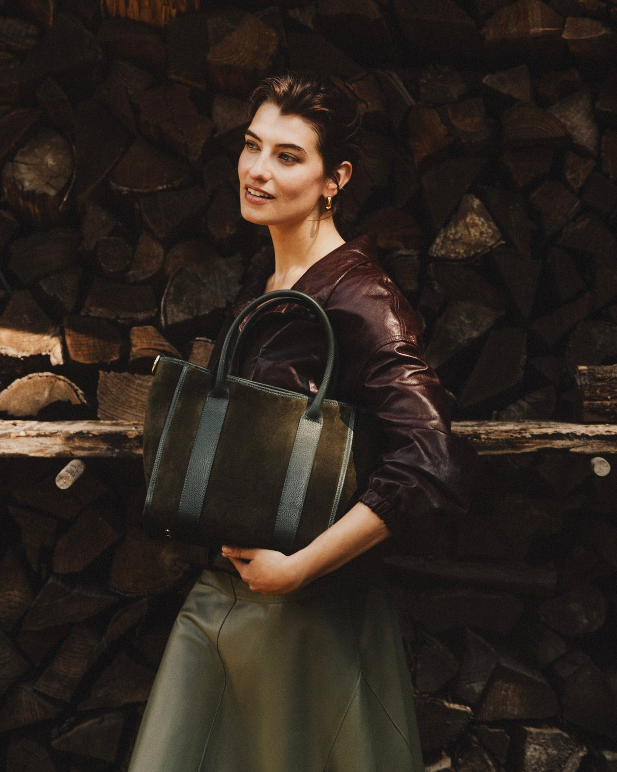 A woman stands in front of a stacked wood wall holding a large structured leather tote bag. She is wearing a dark brown leather top and a light olive green skirt.